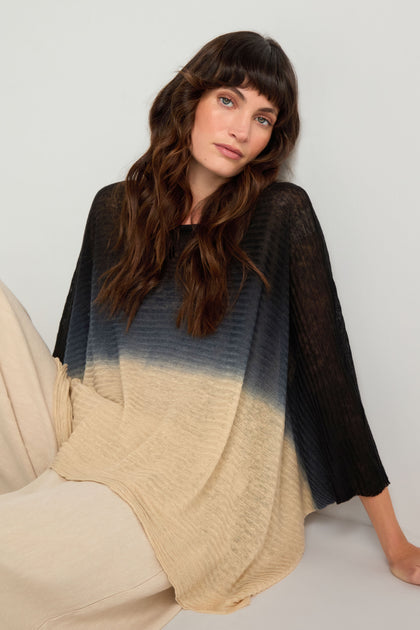 A woman with long brown hair wears the Dip Dye Knit Easy Top in black and beige with light pants, sitting against a light background.