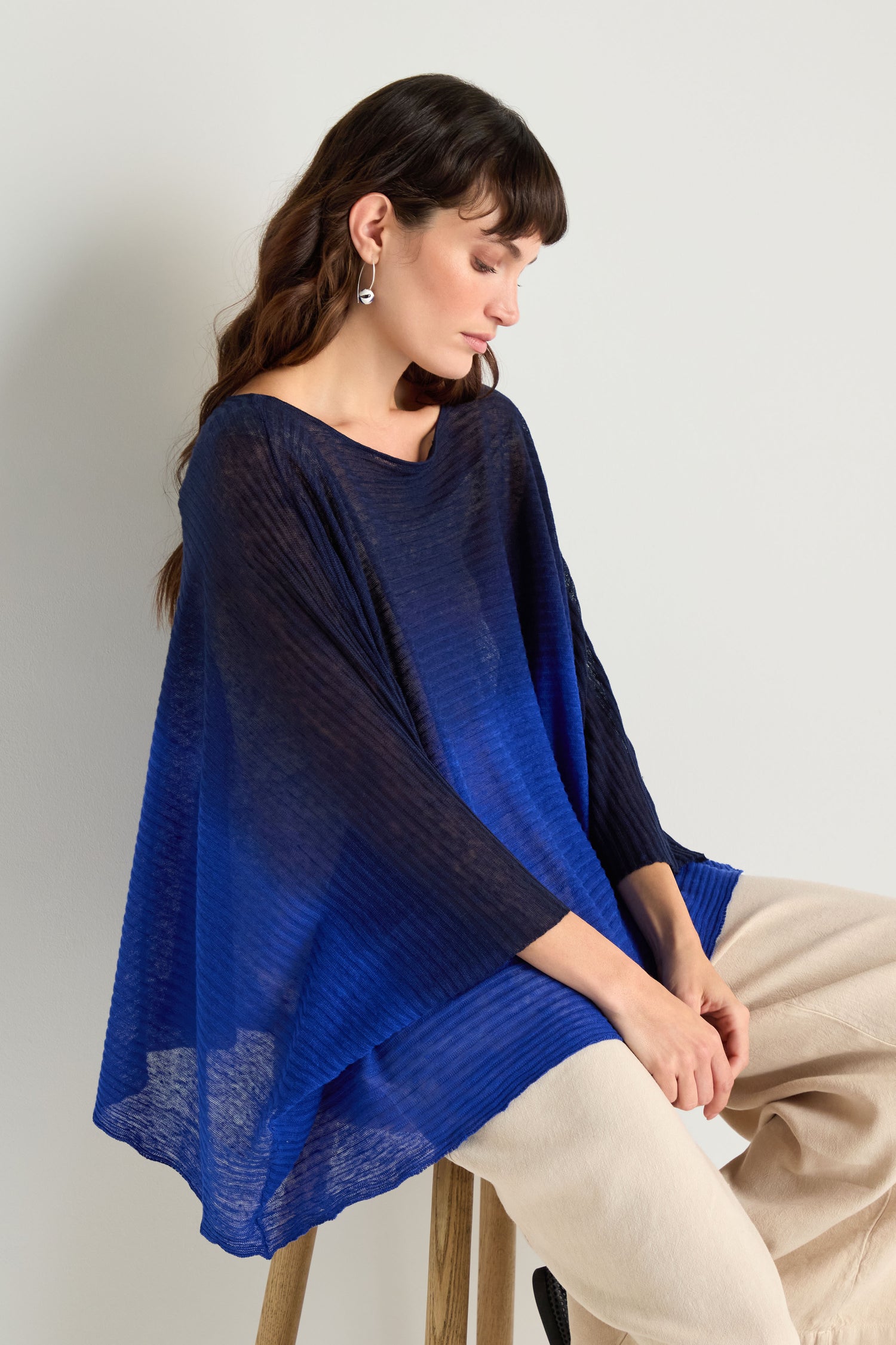 A woman sits on a wooden stool, facing slightly to the side in a relaxed pose, wearing the Navy Blue Dip Dye Knit Easy Top with long sleeves and beige pants against a plain light background.
