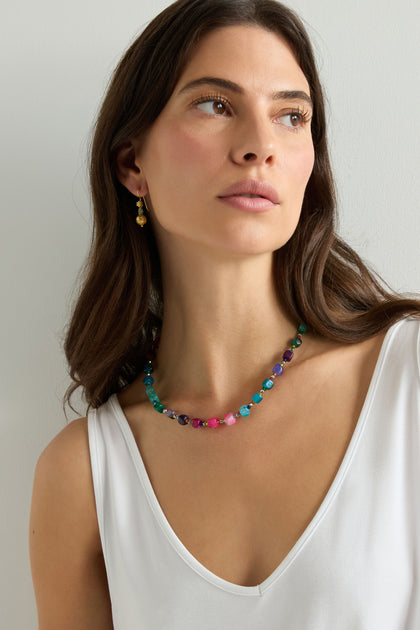 A woman in a sleeveless white top and gold earrings wears the Artisanal Rainbow Agate Cube Necklace, looking slightly to the side against a plain background.