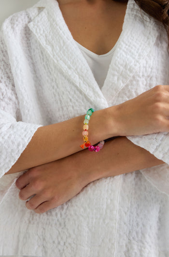 A person in a textured white robe crosses their arms, showcasing the Artisanal Rainbow Agate Bead Bracelet crafted from semi-precious agate beads.