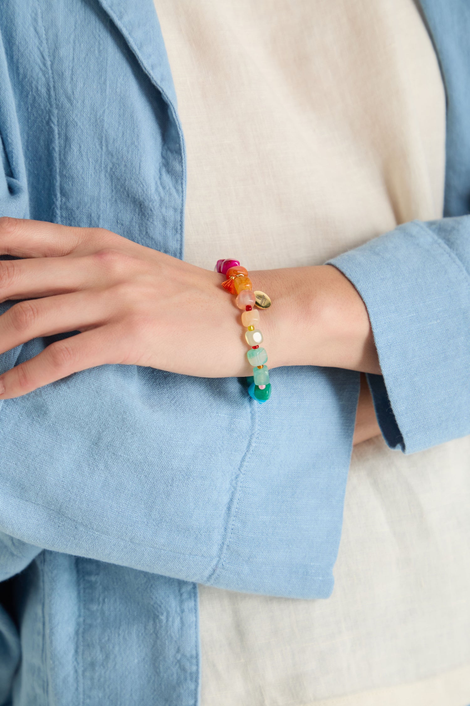 A person in a light blue jacket and cream top showcases the Artisanal Rainbow Agate Bead Bracelet, handmade in Scotland, on their wrist.