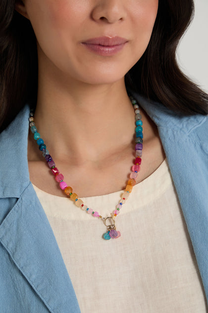 A woman wearing a light blue blazer and cream top showcases the Artisanal Rainbow Agate Toggle Necklace, handmade in Scotland with natural agate stones and charms.