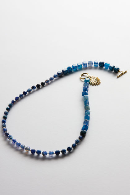 The Artisanal Blue Agate Palm Necklace boasts blue agate stones, a gold toggle clasp, and a delicate gold leaf charm. Handmade in Scotland, this stunning jewellery piece is displayed on a white surface.