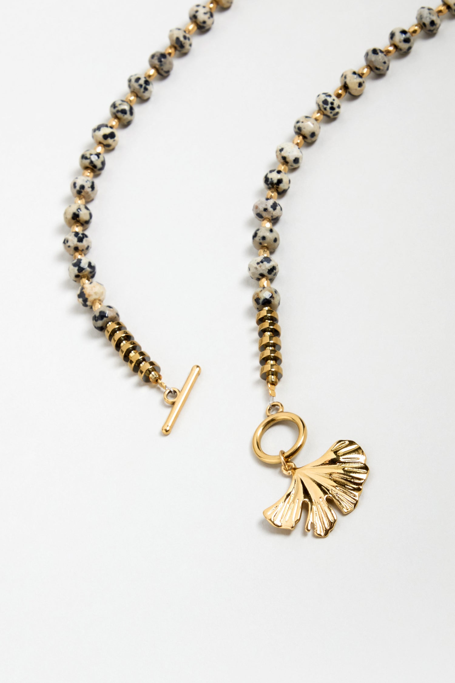 The Dalmatian Stone Ginkgo Leaf Necklace features speckled round Dalmatian stone beads, gold accents, a toggle clasp, and a gold ginkgo leaf pendant, displayed on a white background.
