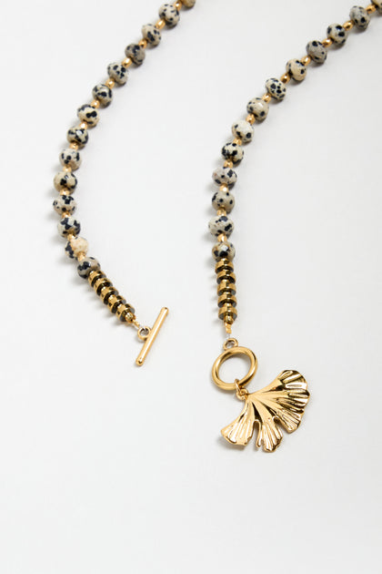 The Dalmatian Stone Ginkgo Leaf Necklace features speckled round Dalmatian stone beads, gold accents, a toggle clasp, and a gold ginkgo leaf pendant, displayed on a white background.