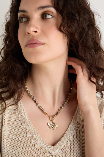 A woman with curly brown hair wears a beige knit top and the Dalmatian Stone Ginkgo Leaf Necklace, looking slightly to the side with one hand near her neck.