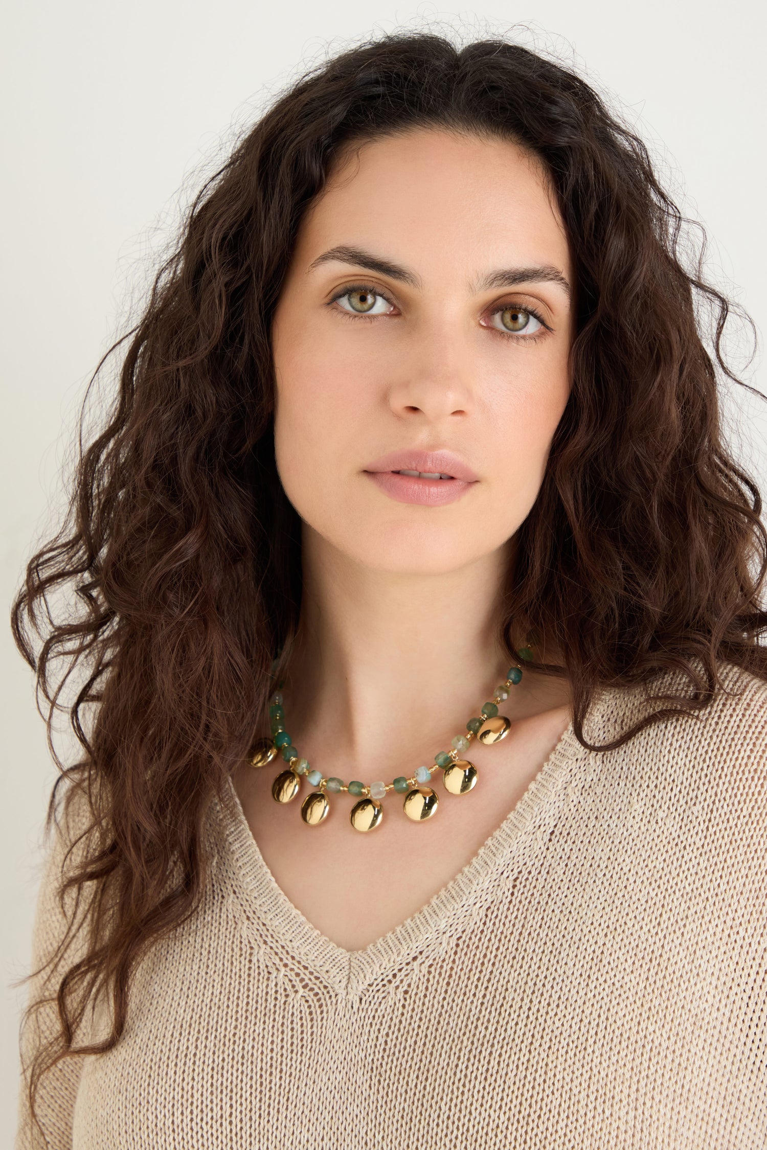 A woman with long curly brown hair wears a beige knit top and the Faceted Green Agate Charm Necklace, looking at the camera against a plain background.