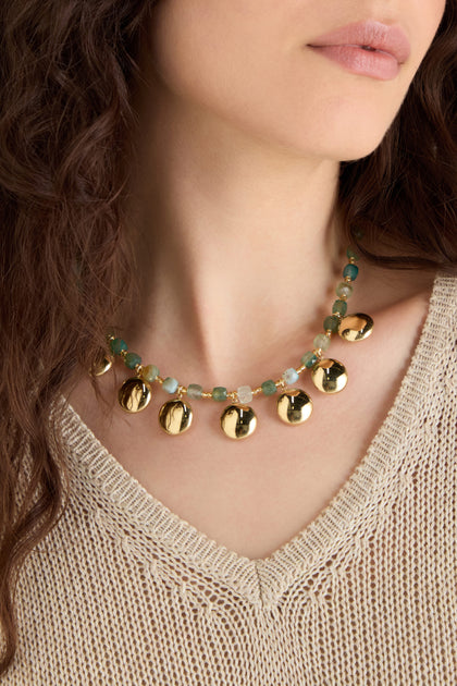 A woman wearing a beige knit top and the Faceted Green Agate Charm Necklace, featuring large gold pendants and green beads.