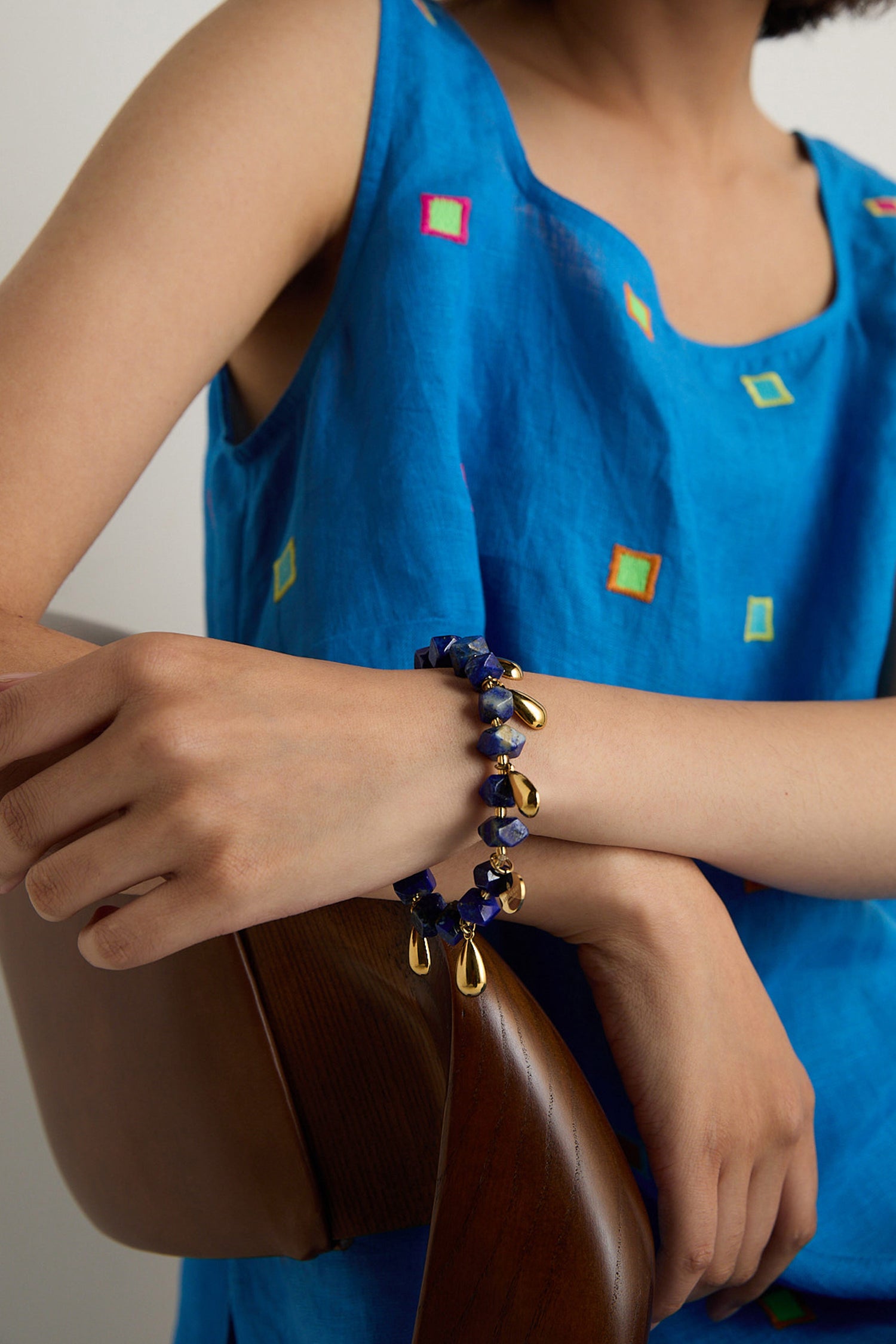 A person in a blue sleeveless top with colorful squares rests an arm on a wooden chair, wearing the handmade Lapis Lazuli Teardrop Bracelet, crafted in Scotland.