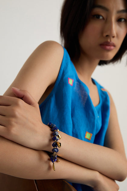 A person in a sleeveless blue top with colorful square accents and folded arms showcases the handmade Lapis Lazuli Teardrop Bracelet from Scotland.