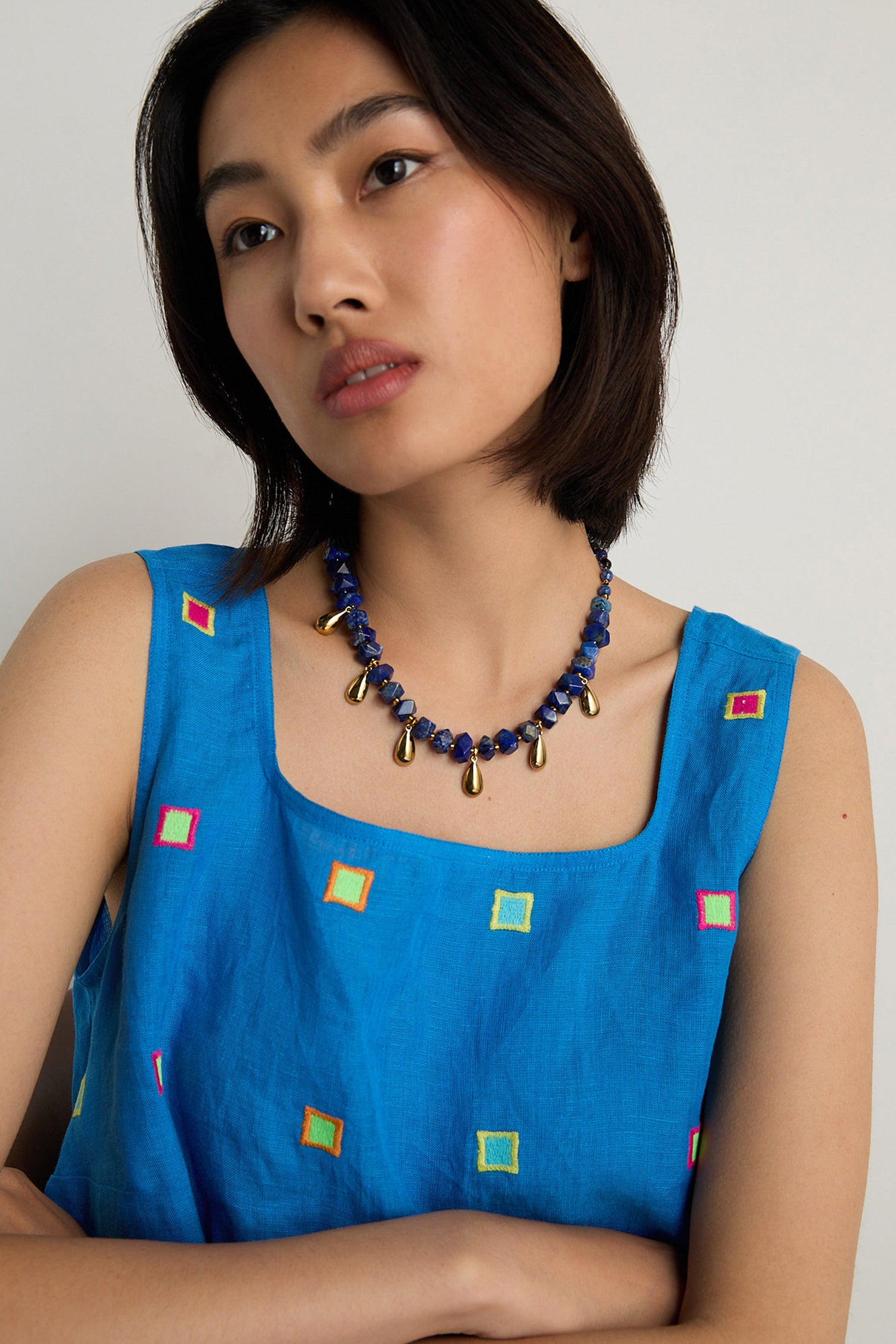 A woman with short dark hair wears a blue sleeveless top with colorful squares and a handmade Lapis Lazuli Teardrop Necklace from Scotland, featuring bold blue and gold beads.