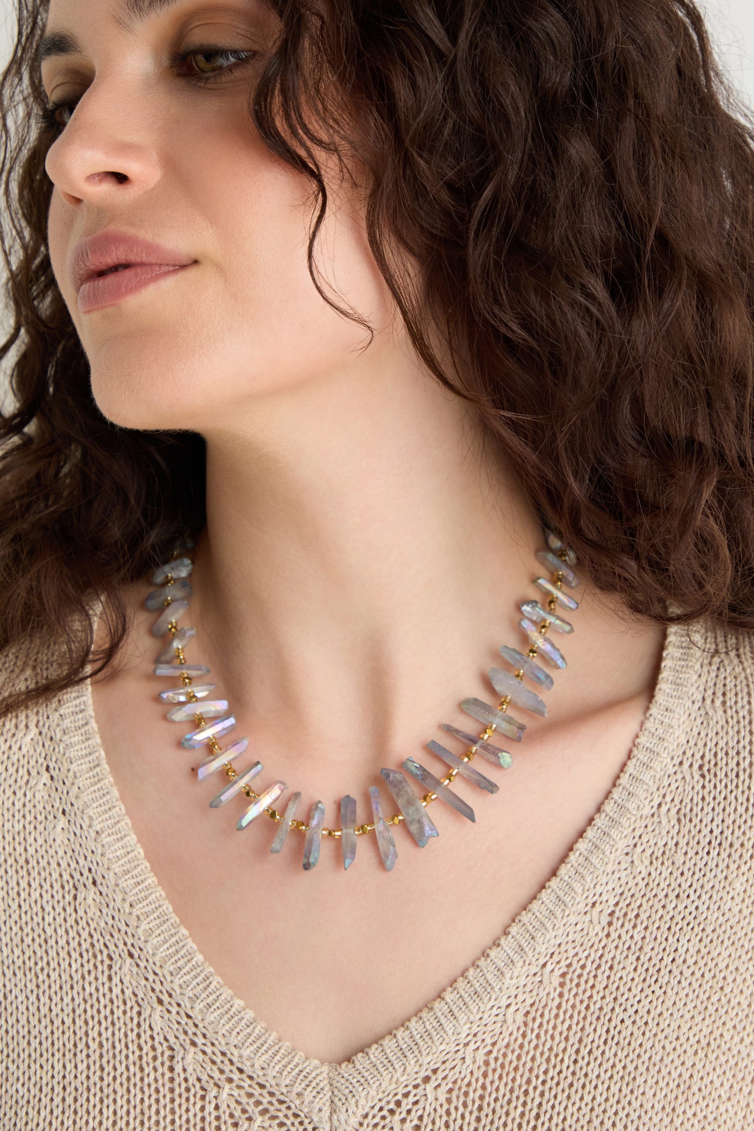 A woman with curly brown hair wears a beige knit top and the Slate Quartz Necklace, a gold-plated statement piece featuring irregularly shaped, light-colored stones and gold accents.