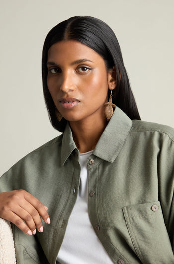 A person with long hair wears an olive green jacket and hand crocheted, snowdrop-shaped earrings, sitting against a light background.