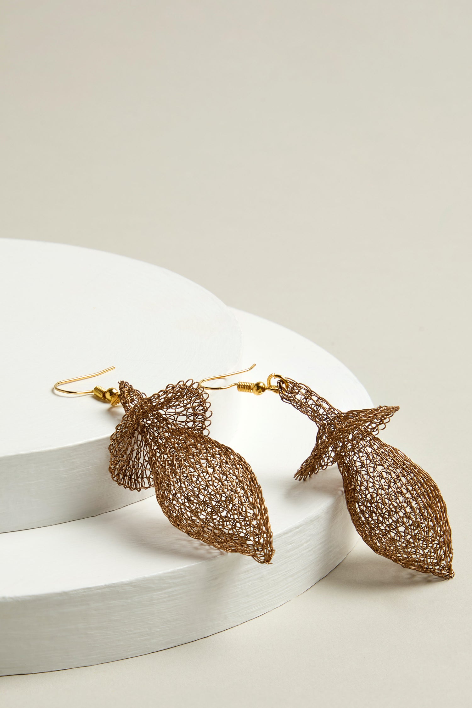 The Hand Crocheted Snowdrop Earrings, featuring exquisite metallic crochet work in a teardrop shape, are elegantly displayed on round white platforms, with their gold hooks adding a touch of sophistication.