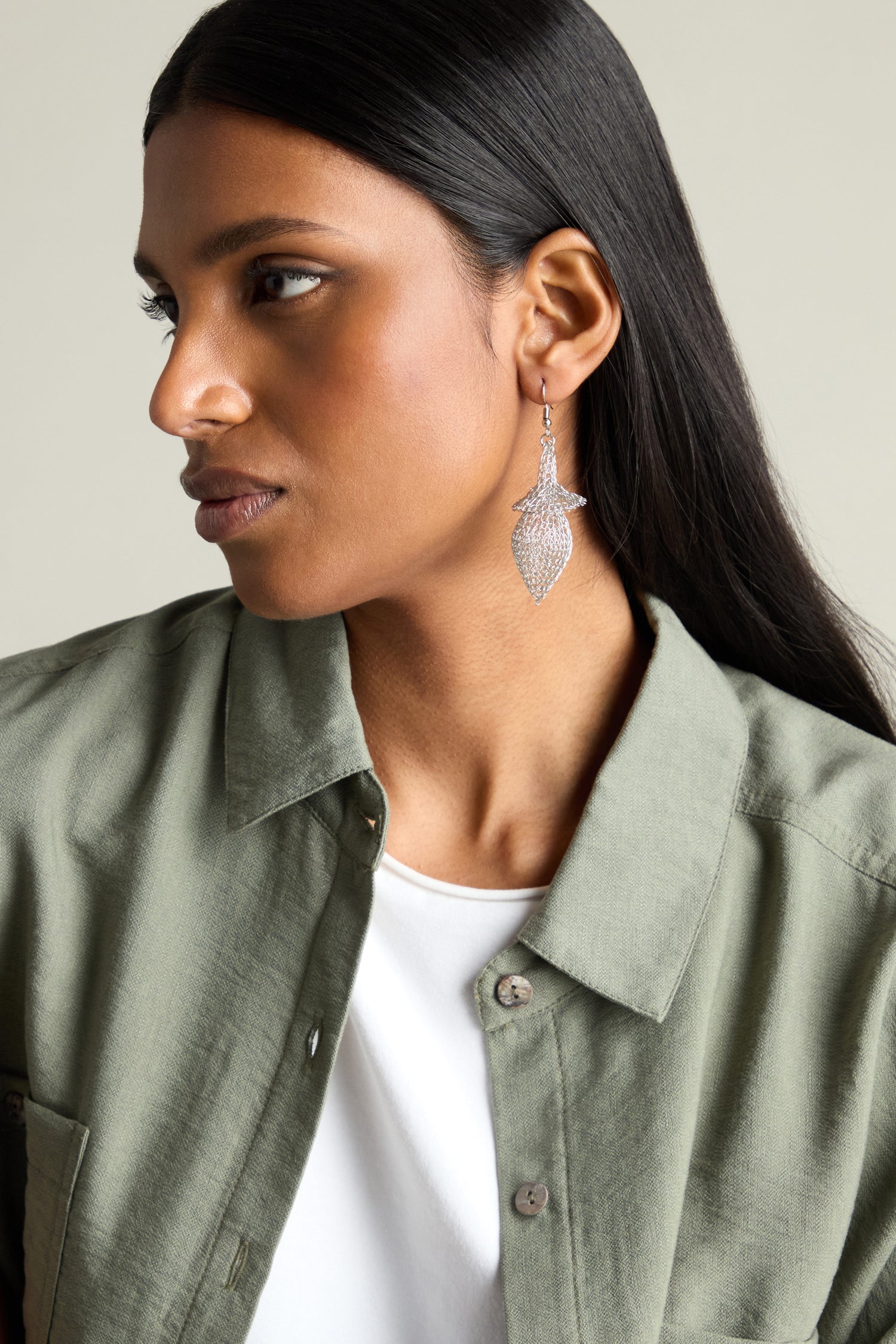 A woman with long black hair wears a green button-up shirt and a large, hand-crocheted Snowdrop earring, looking to the side.