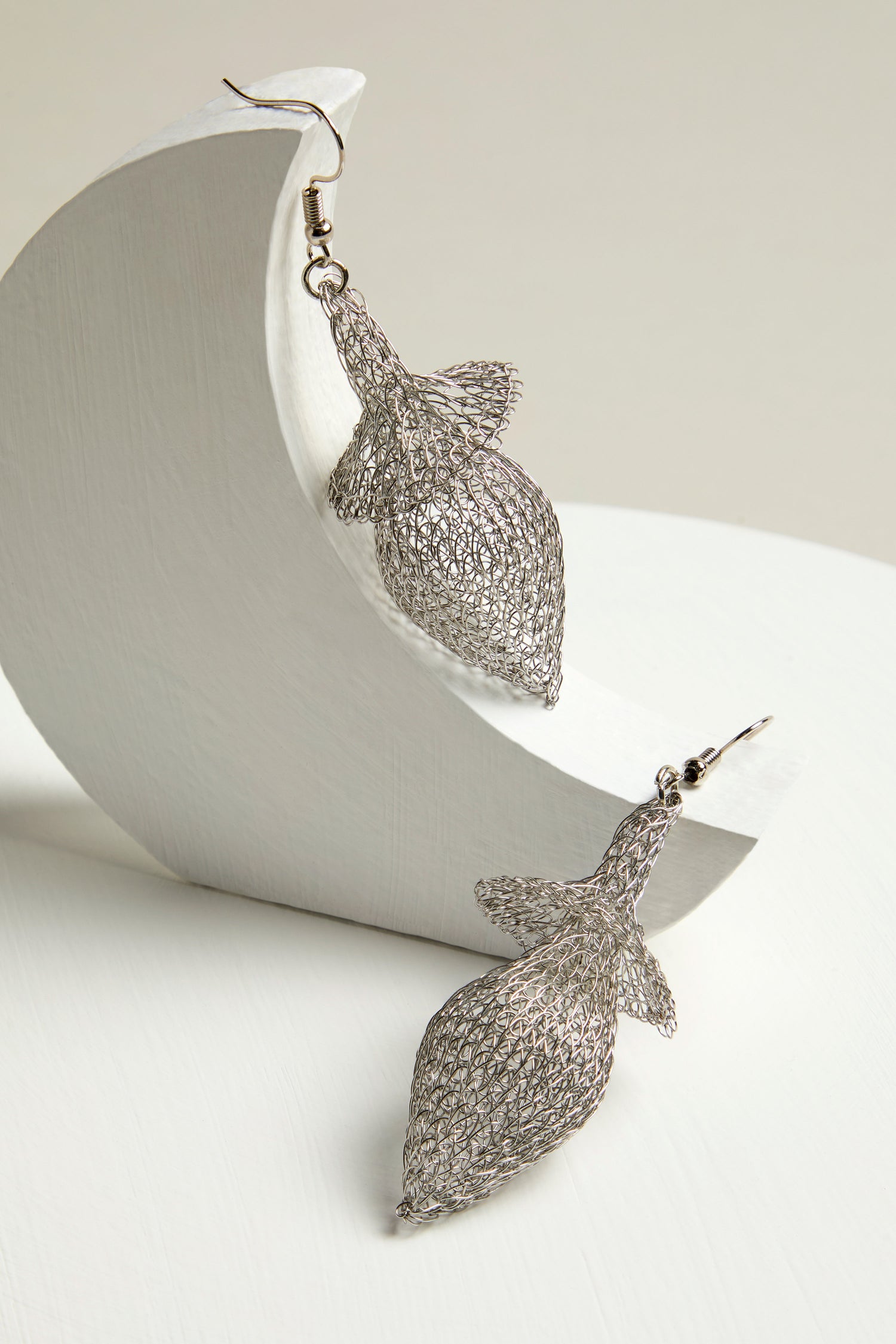 Two intricately designed Hand Crocheted Snowdrop Earrings, shaped like silver fish, are elegantly displayed on a curved white stand against a neutral background.