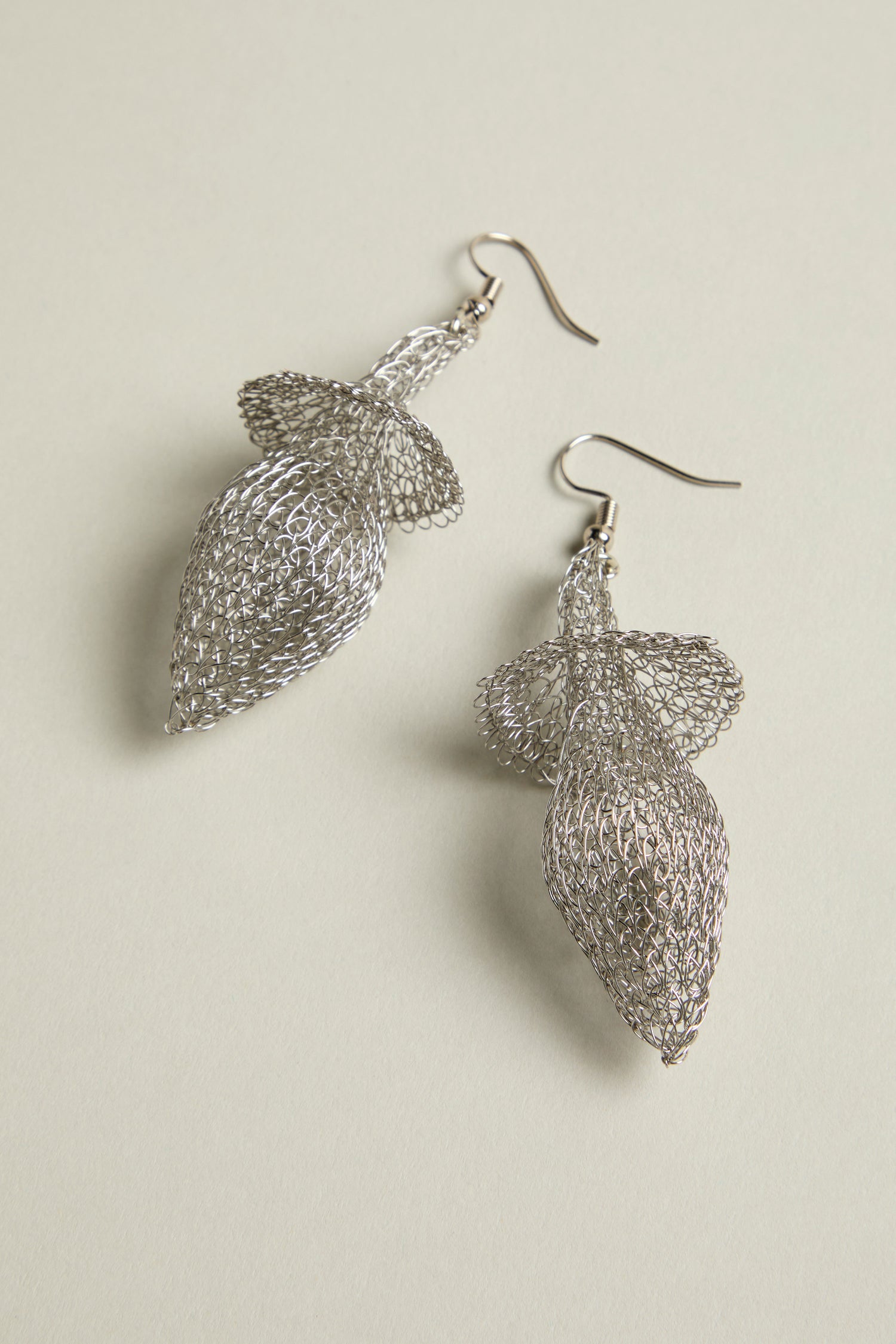 The Hand Crocheted Snowdrop Earrings are intricately crafted from silver wire mesh, showcasing a delicate leaf-like design against a light background.