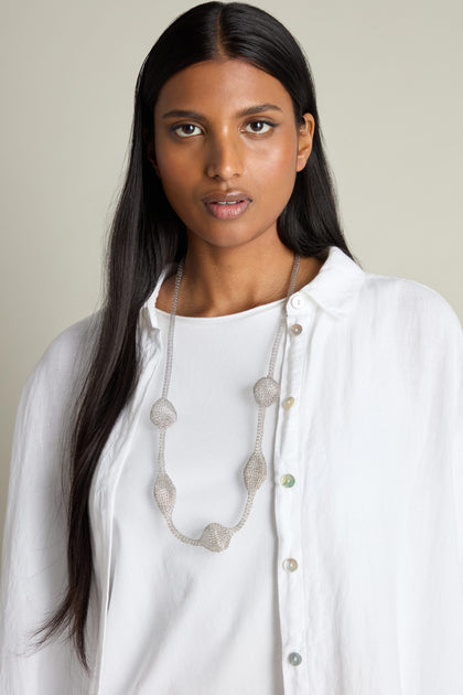 A person with long black hair, wearing a white shirt and the Hand Crocheted Orb Necklace, looks directly at the camera.