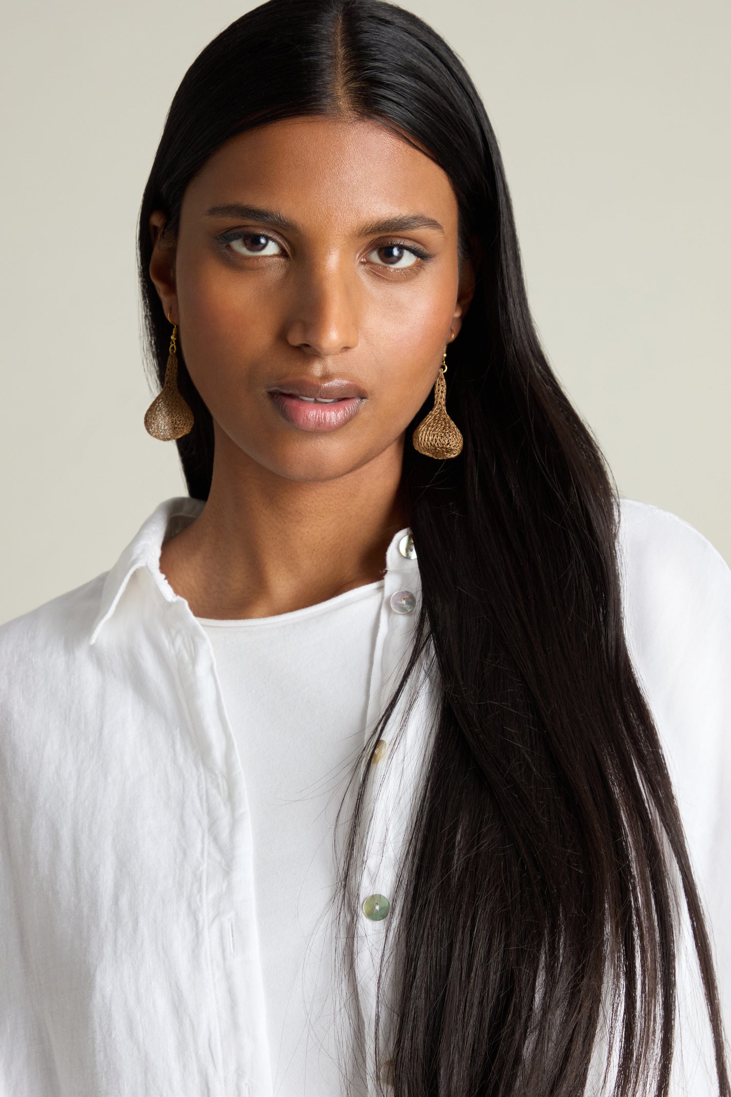 A person with long dark hair in a white shirt poses gracefully against a plain backdrop, highlighting the Hand Crocheted Teardrop Earrings. These earrings, expertly crafted by artisans, shimmer delicately with metallic threads.