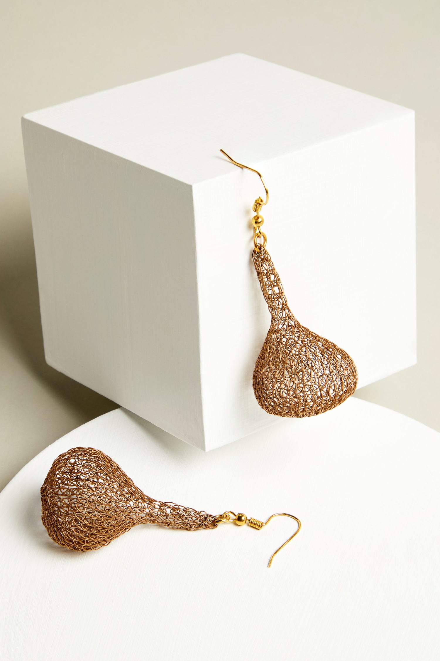 A pair of Hand Crocheted Teardrop Earrings featuring gold mesh elegantly frame and cradle a white cube, highlighting their artisanal craftsmanship and glimmering metallic threads.