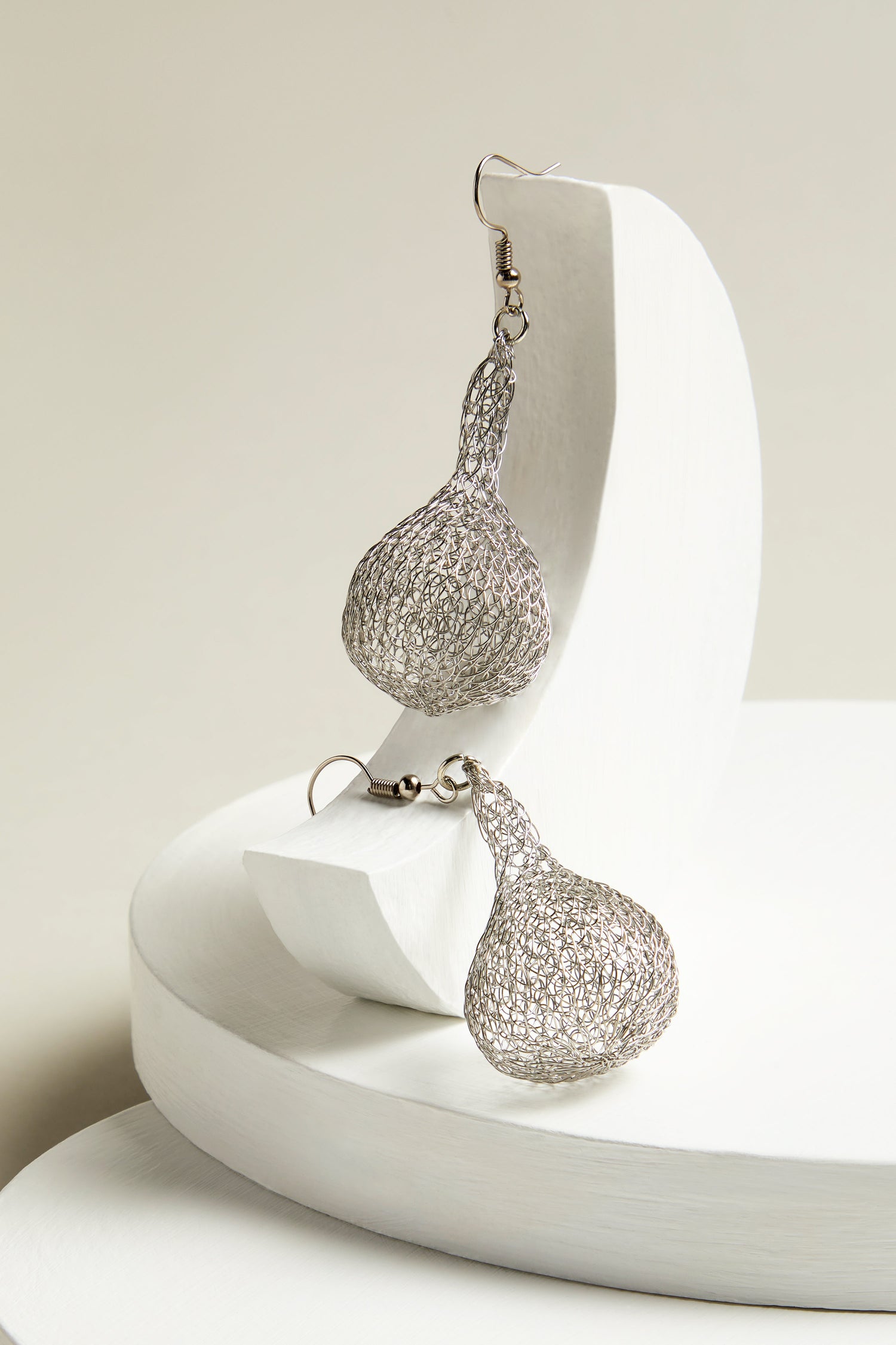 The Hand Crocheted Teardrop Earrings, made from silver wire mesh, are elegantly presented on a white curved surface, highlighting their exquisite artisanal craftsmanship.