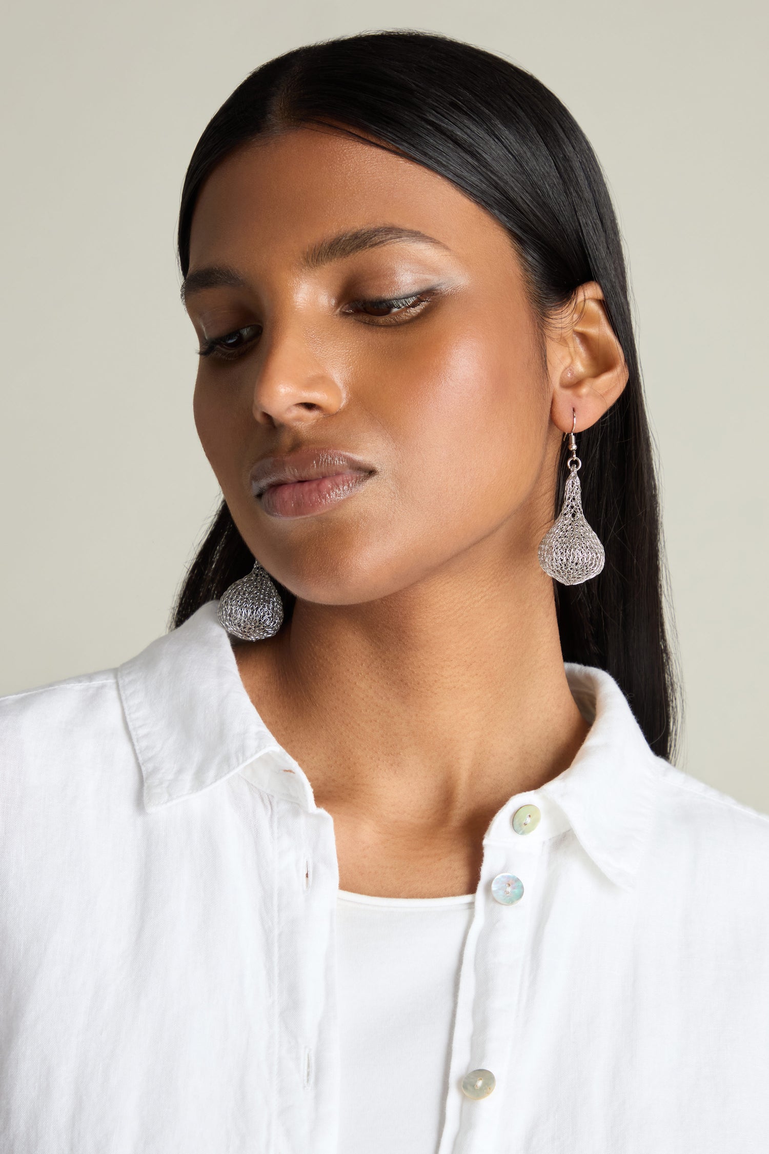 A person with long black hair wearing a white shirt and Hand Crocheted Teardrop Earrings, displaying exceptional artisanal craftsmanship, gazes downward.