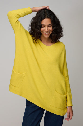 A woman with curly hair smiles and lifts an arm, wearing the Two Pocket Oversize Knit in yellow from Amazing Woman, styled with dark pants against a simple backdrop.