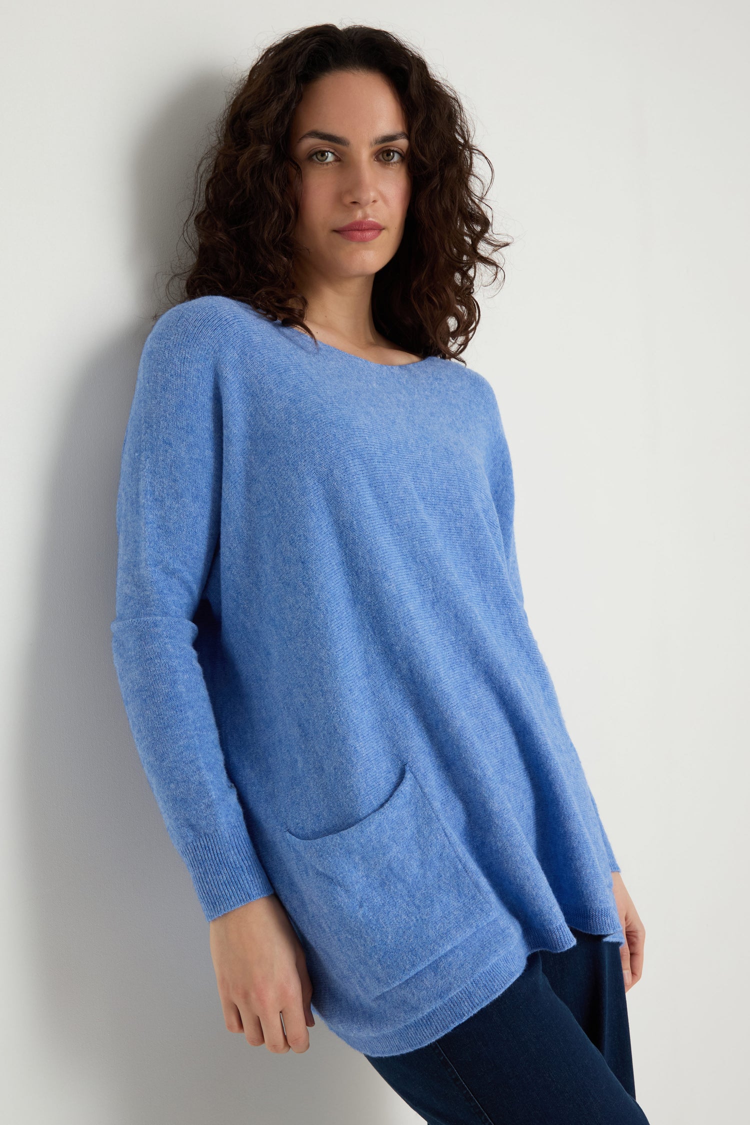 A person with curly brown hair wears the Two Pocket Oversize Knit—a light blue, oversized women's knitwear jumper with front pockets—and dark pants, standing against a white wall.