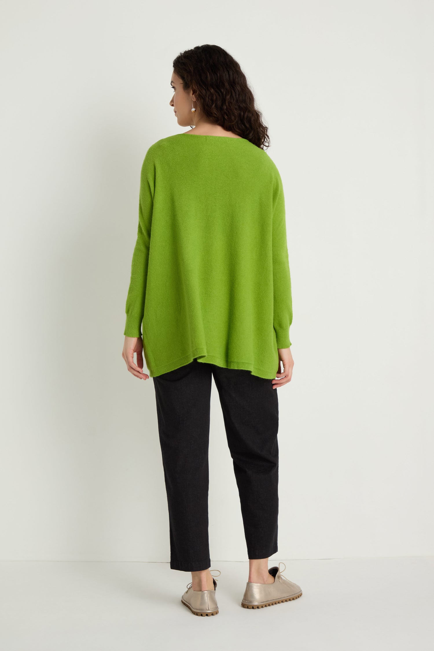 A person stands against a white wall, facing away, wearing the Two Pocket Oversize Knit in green from women's knitwear, paired with black pants and beige shoes.