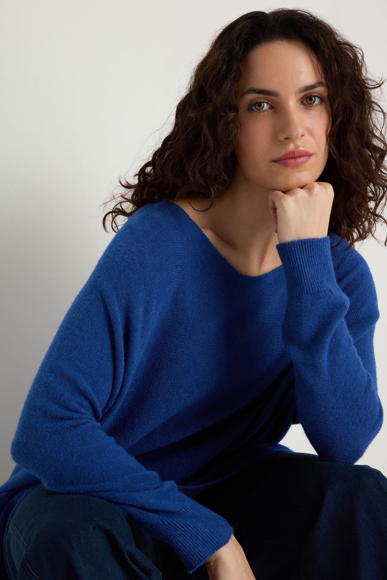 A woman with wavy brown hair wears the Two Pocket Oversize Knit, sitting with her chin on her hand and looking at the camera against a plain light background.