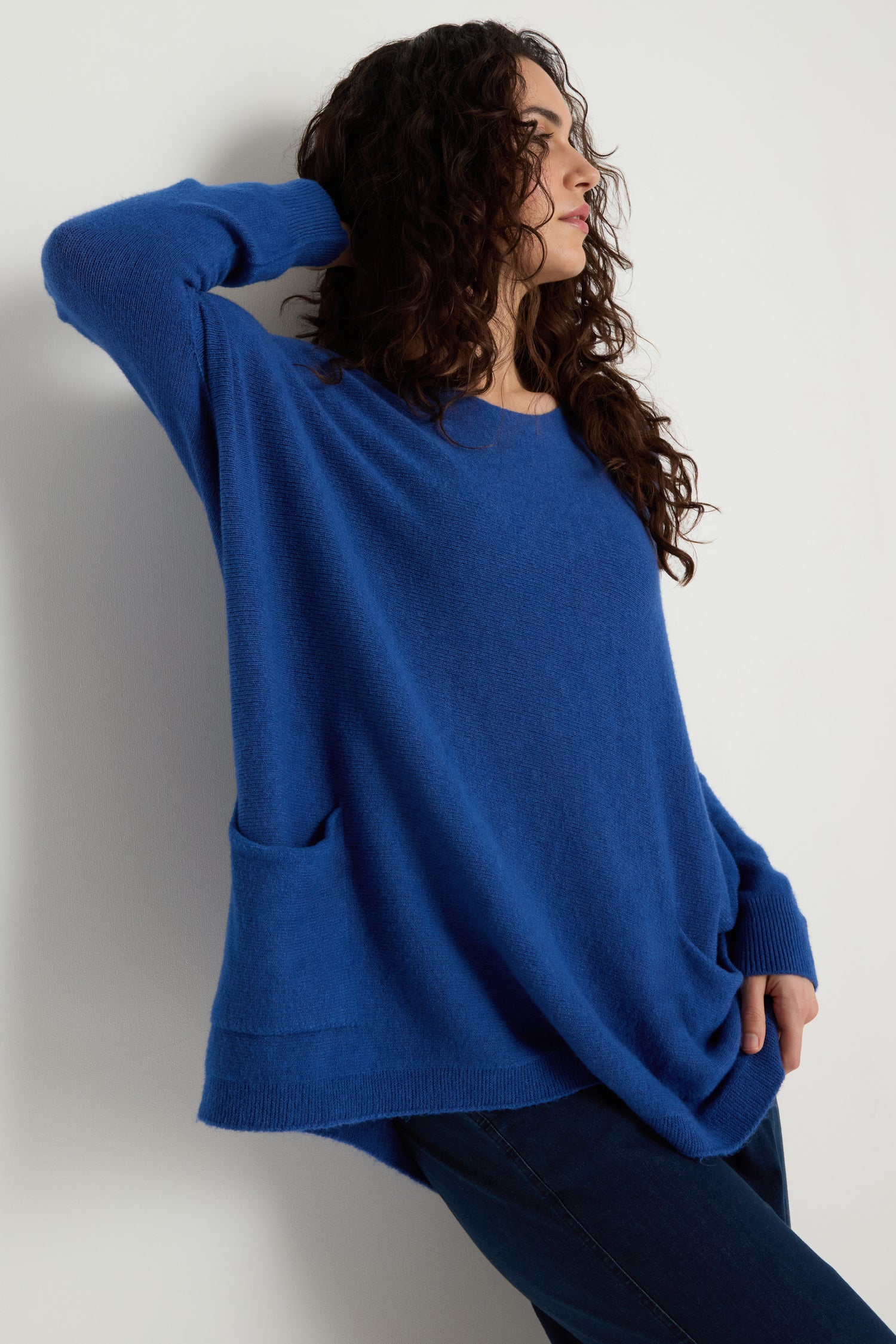 A woman with curly hair wears the blue Two Pocket Oversize Knit and dark jeans, standing against a white wall and looking to the side with one hand behind her head.