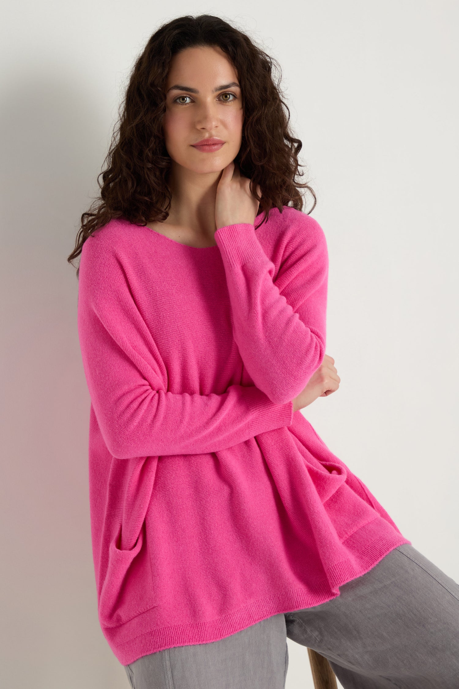 A person with long curly hair sits on a stool against a plain white background, wearing the Two Pocket Oversize Knit in pink and light gray pants.