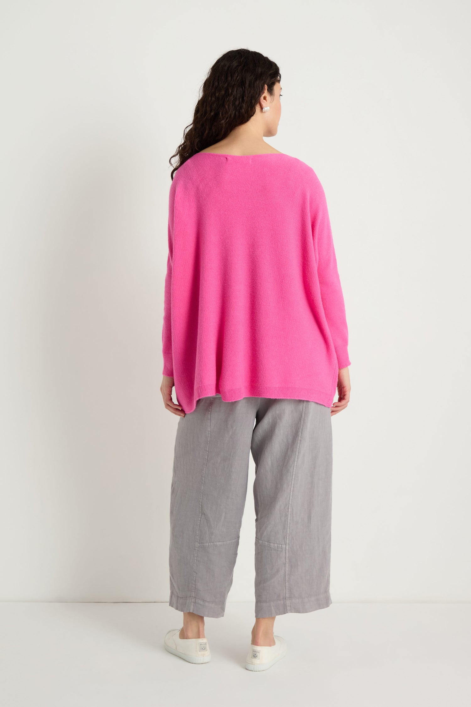 Someone stands facing away in a bright pink Two Pocket Oversize Knit sweater, paired with gray wide-leg pants and white shoes, set against a plain white background.
