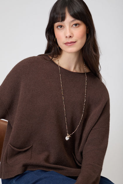 Woman with long dark hair wears the Two Pocket Knit jumper and a long necklace, seated and looking at the camera against a plain background.