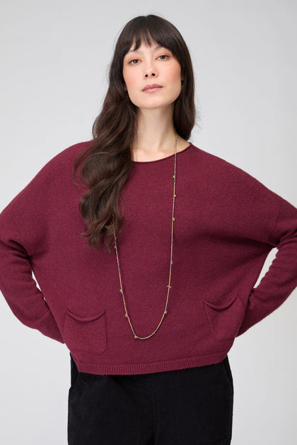 A woman with long brown hair stands against a plain light background, wearing the maroon Two Pocket Knit jumper with front pockets, black pants, and a long necklace.