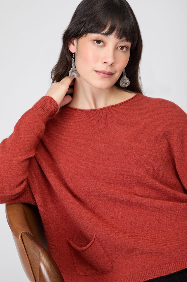 A woman with dark hair, wearing silver earrings and the rust-colored Two Pocket Knit jumper, sits comfortably on a brown chair.