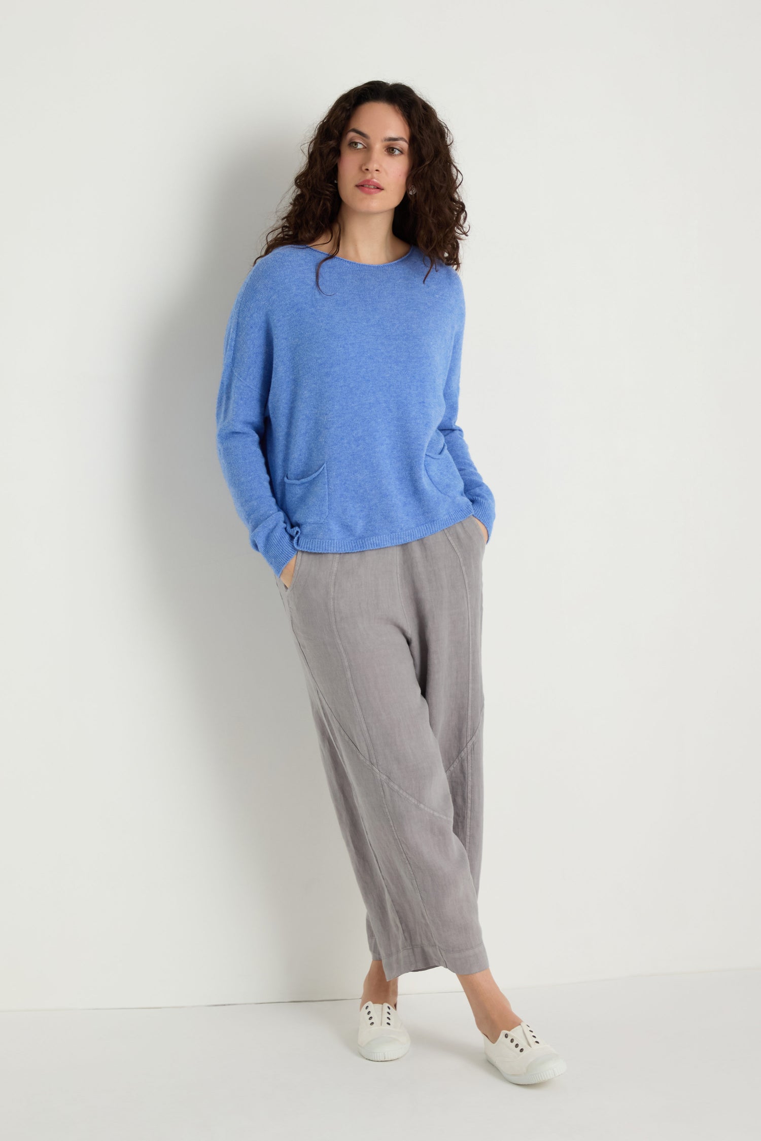 A woman stands against a plain white wall, wearing the Two Pocket Knit in blue, loose grey pants, and white sneakers. With hands in her pockets and looking aside, this sweater is perfect as a versatile layering piece.