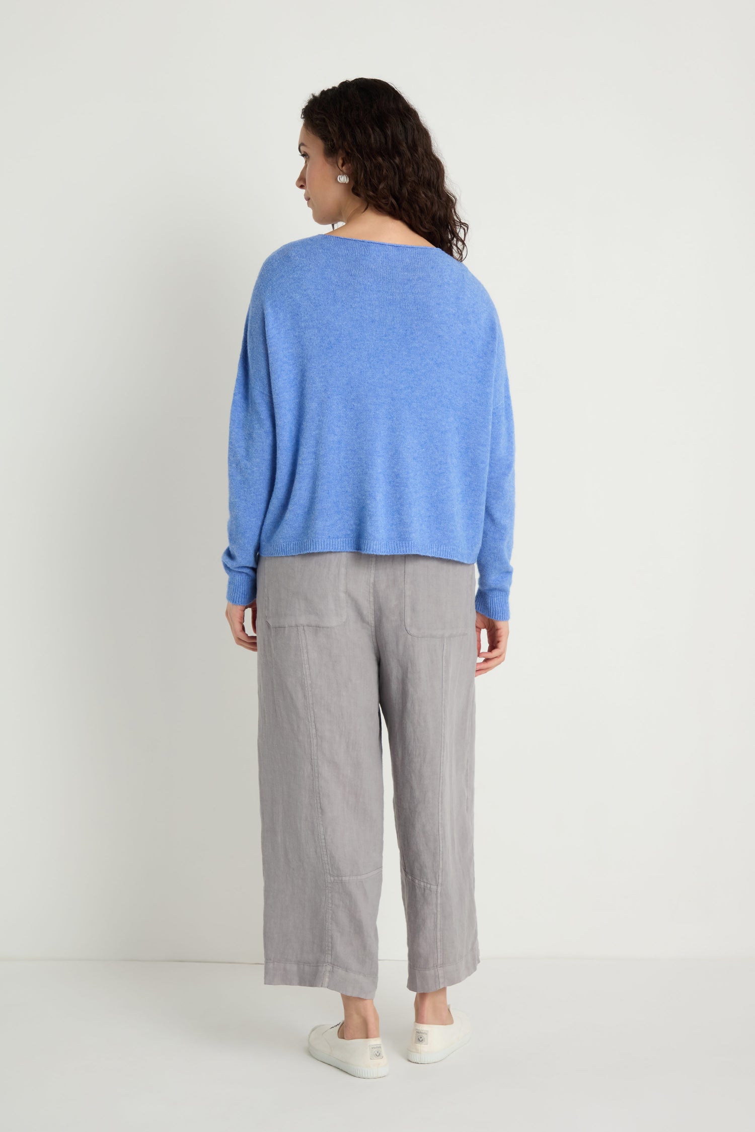 A person stands facing away, wearing the Two Pocket Knit sweater—perfect for layering every day—paired with gray wide-leg pants and white slip-on shoes against a plain white background.