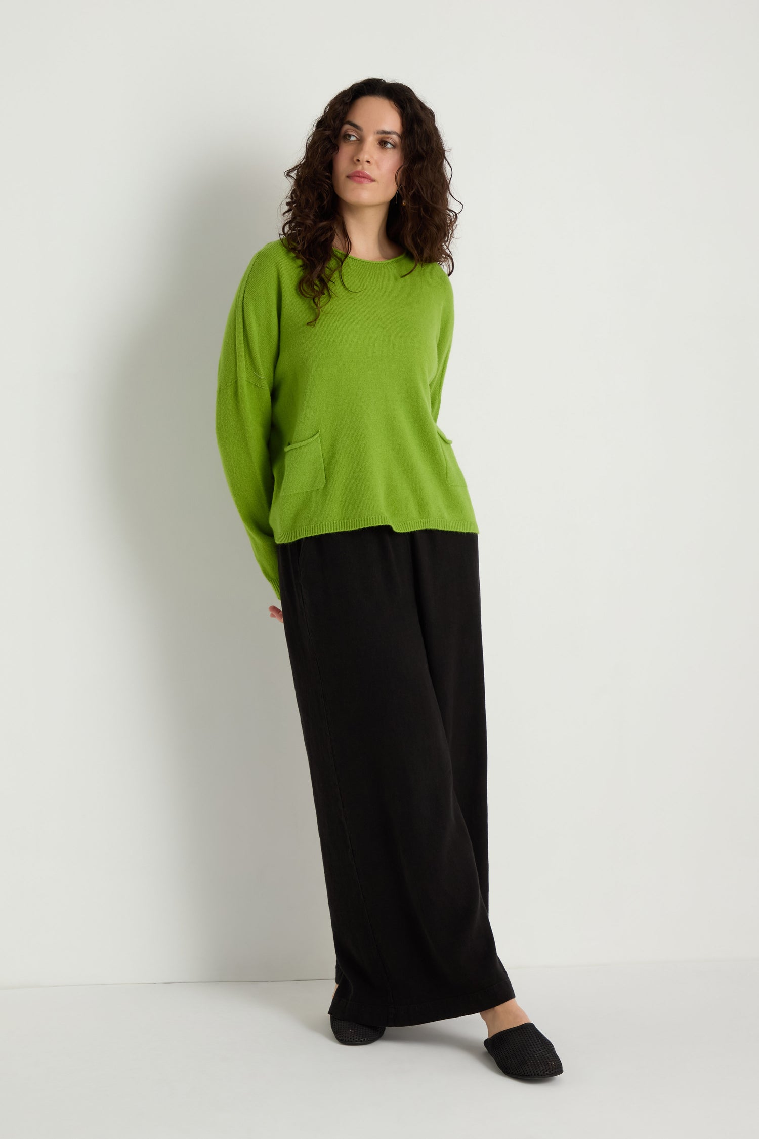 A person stands against a white wall wearing the green Two Pocket Knit sweater, ideal for layering in daily outfits, paired with loose black pants and black slippers.