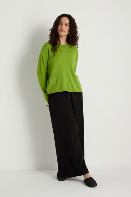 A person stands against a white wall wearing the green Two Pocket Knit sweater, ideal for layering in daily outfits, paired with loose black pants and black slippers.