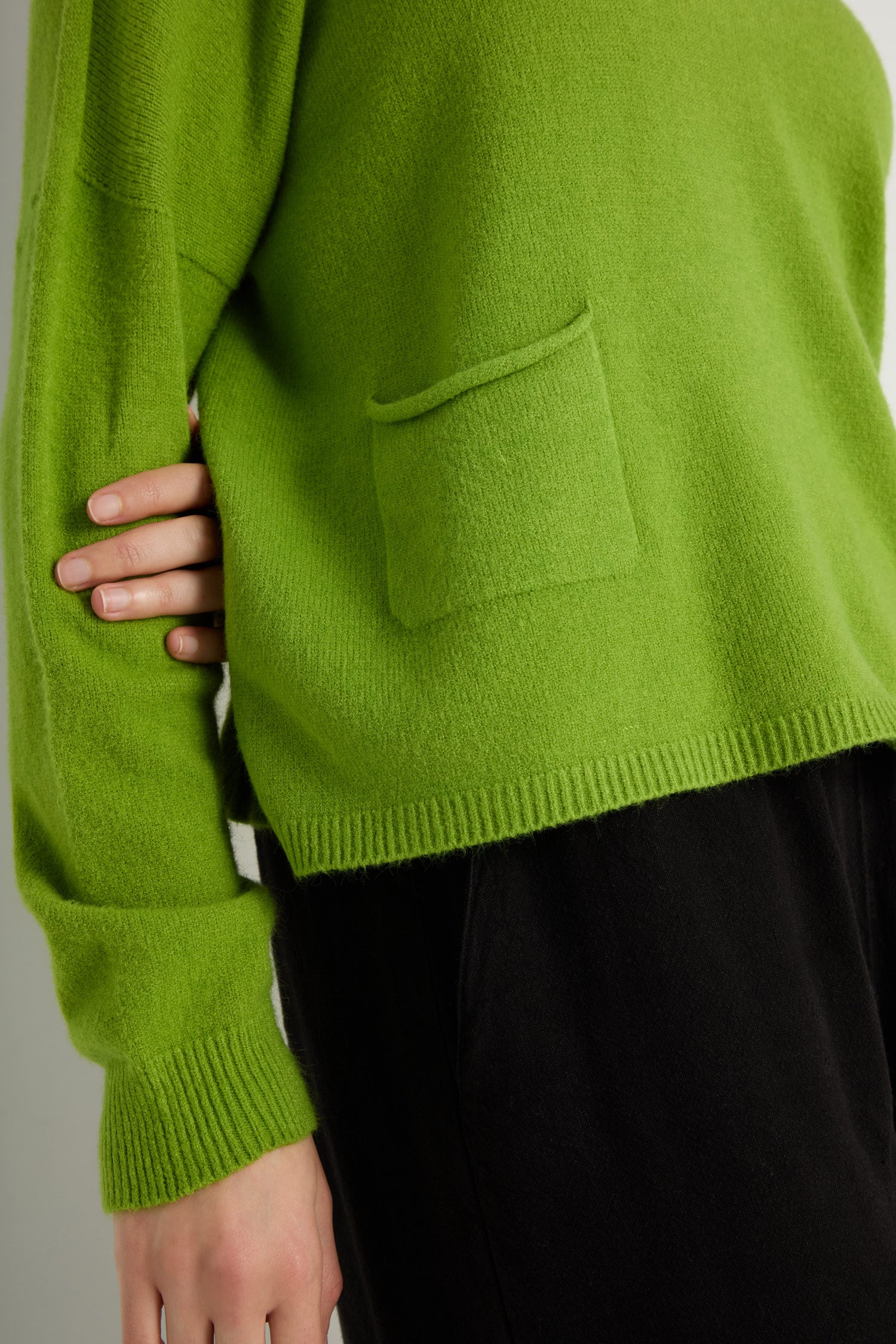 Person wearing a vibrant green Two Pocket Knit sweater and black pants, one hand at their side—an ideal everyday layering piece.