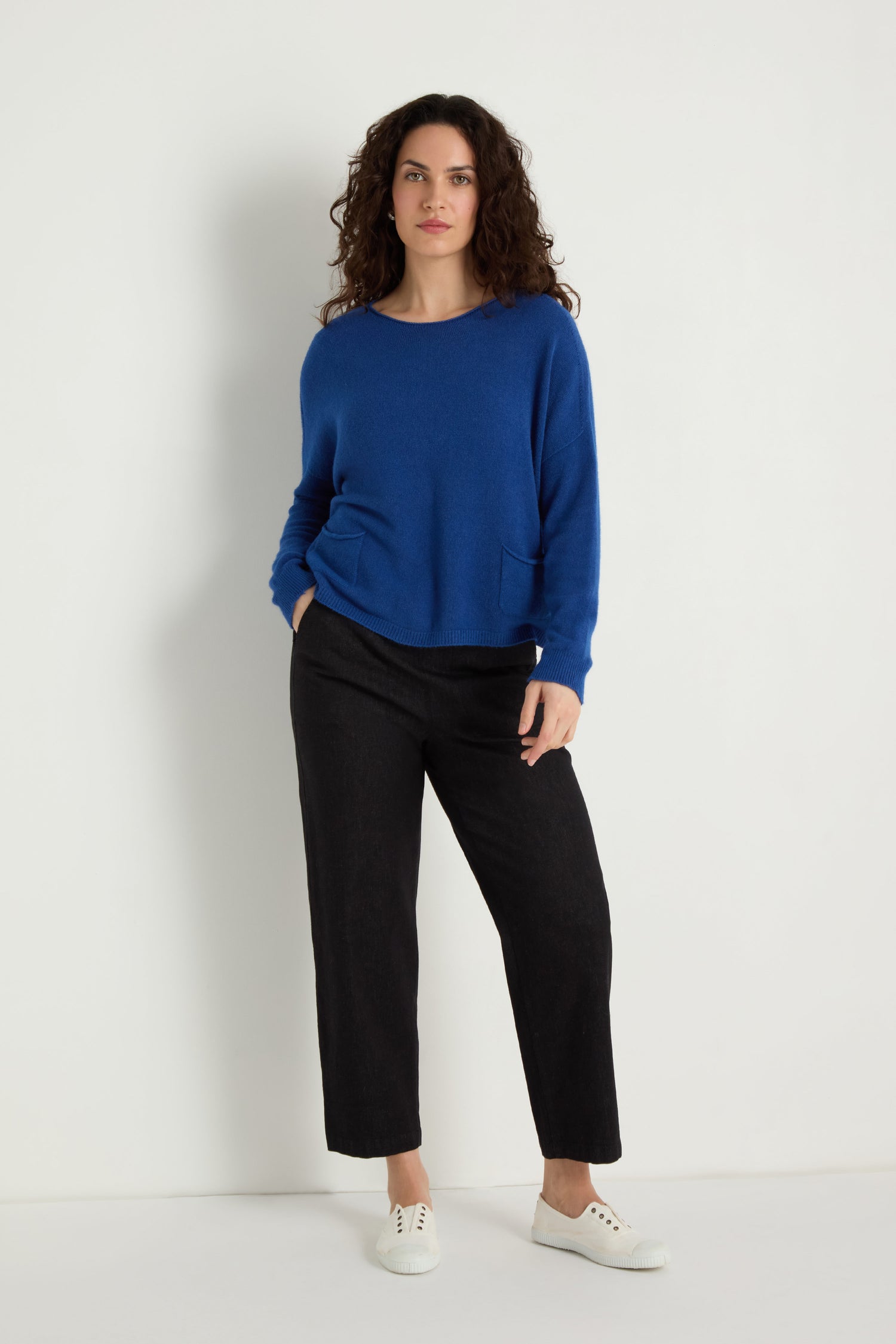 A woman stands against a white wall wearing the Two Pocket Knit—a blue women's sweater perfect for layering—paired with black pants and white sneakers, one hand in her pocket.