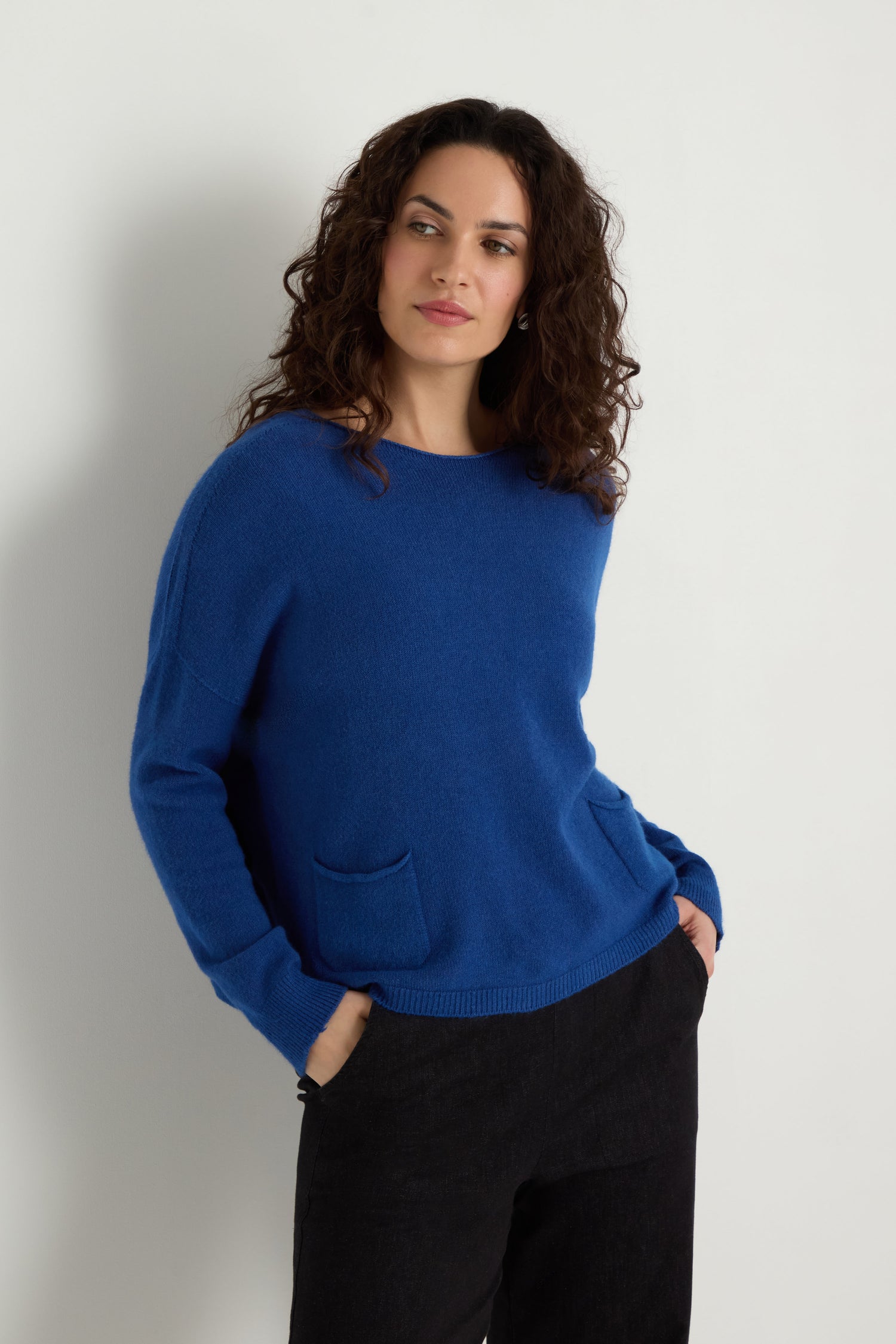 Woman with curly hair wears the Two Pocket Knit in blue with black pants, standing against a plain white background and hands in pockets—an ideal everyday layering piece.