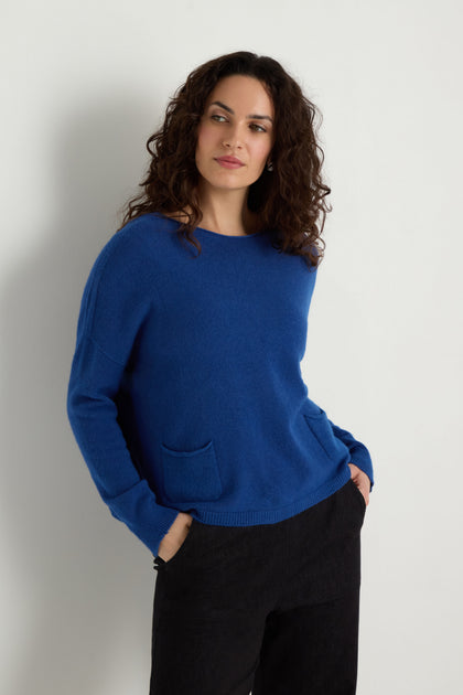 Woman with curly hair wears the Two Pocket Knit in blue with black pants, standing against a plain white background and hands in pockets—an ideal everyday layering piece.