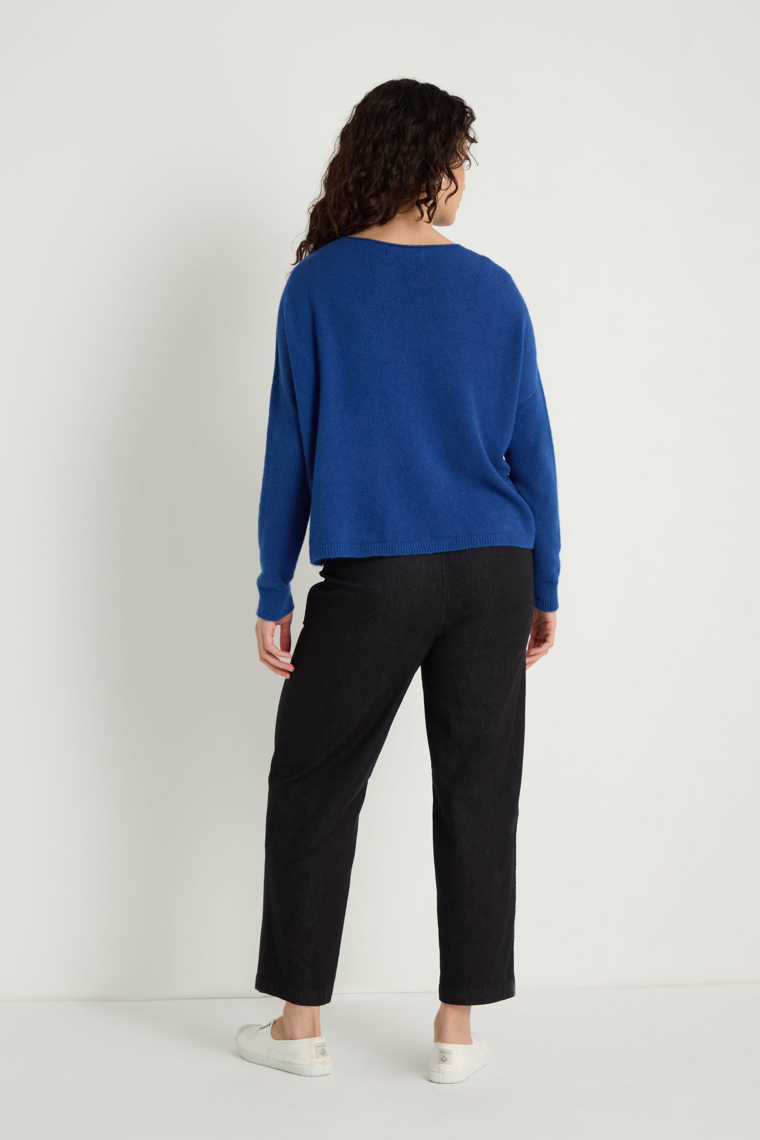 Facing away, a person with curly hair wears the Two Pocket Knit in blue—perfect for layering daily—with black pants and white shoes, against a plain white background.