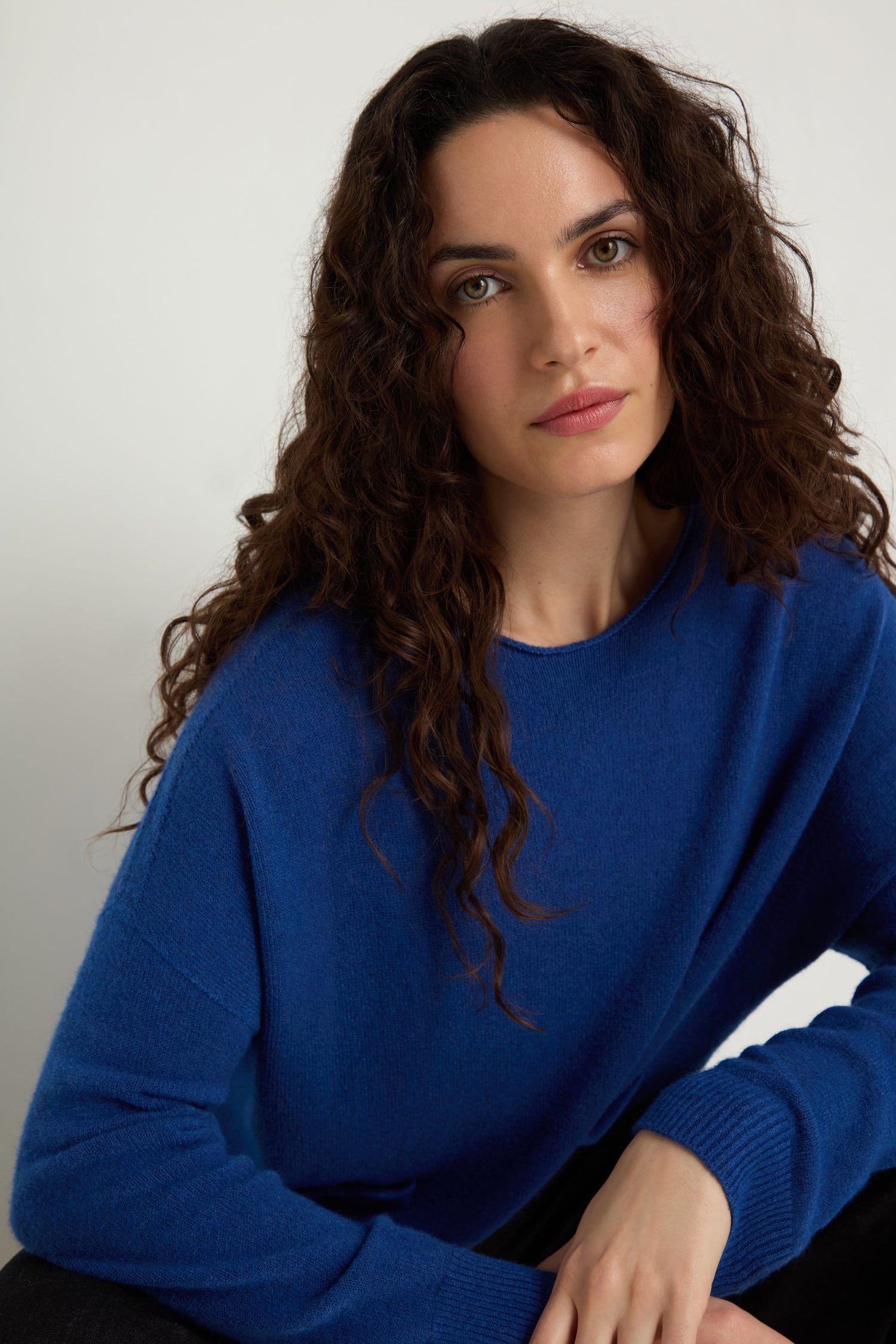 A woman with long curly brown hair wears the Two Pocket Knit in blue, sitting against a plain background and looking directly at the camera with a neutral expression.