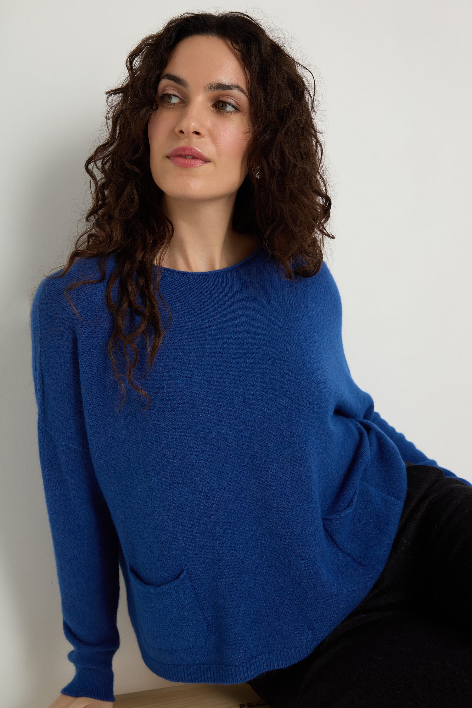 A woman with long curly hair wears the Two Pocket Knit sweater and black pants, sitting against a plain white background—an ideal layering piece for everyday wear.