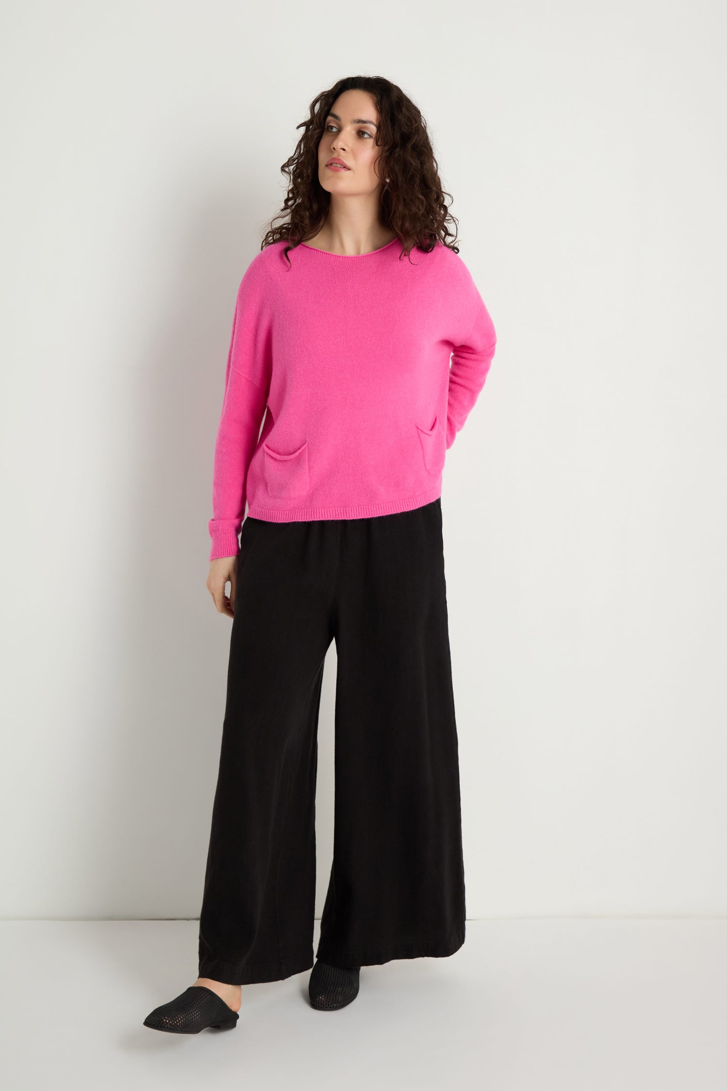 A person stands against a white wall, wearing the bright pink Two Pocket Knit sweater—an ideal everyday layering piece—with wide-leg black pants and black slip-on shoes.