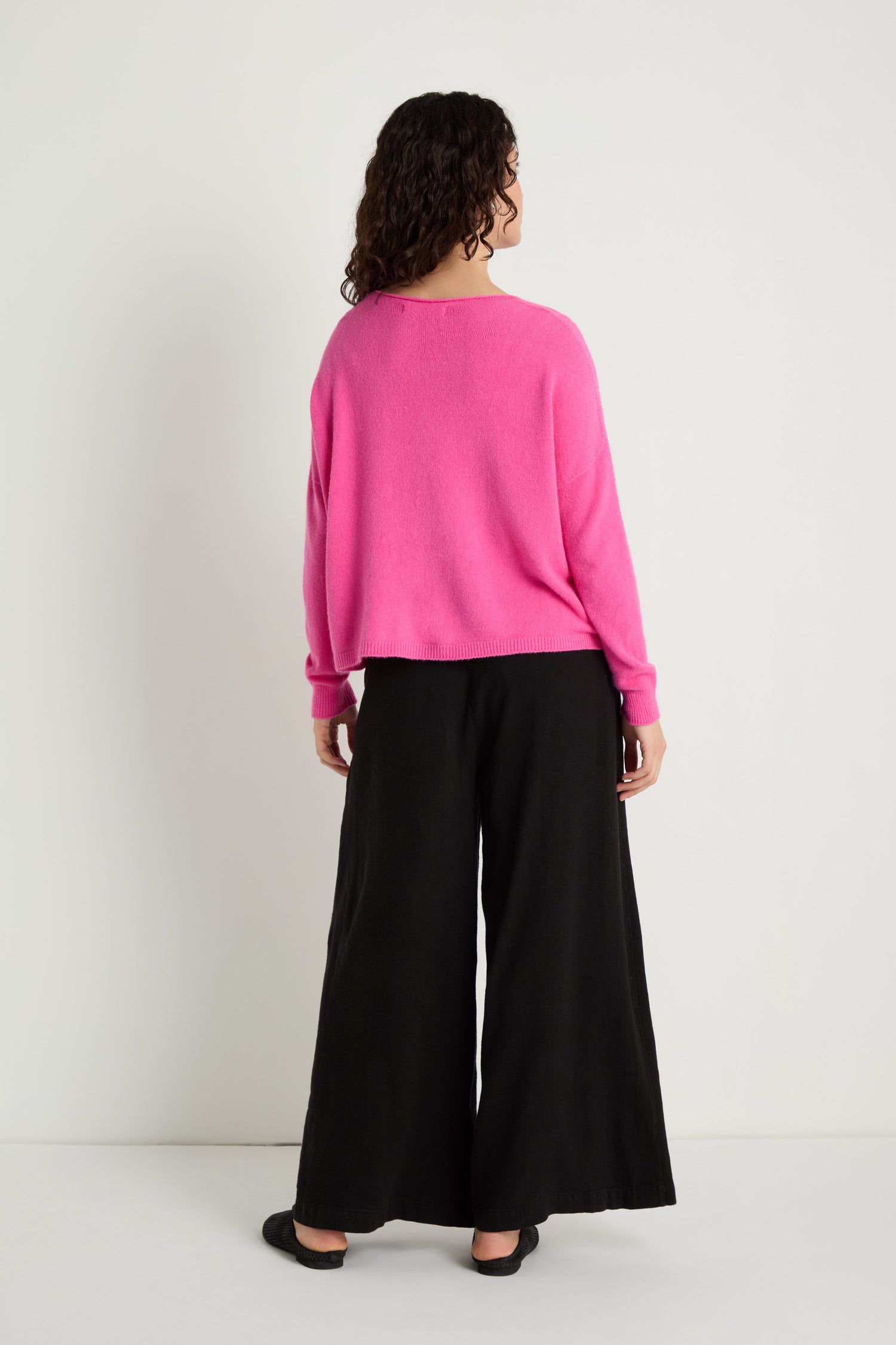 Model stands facing away, wearing the Two Pocket Knit in bright pink—ideal for everyday layering—paired with black wide-leg pants and black shoes, all set against a plain white background.