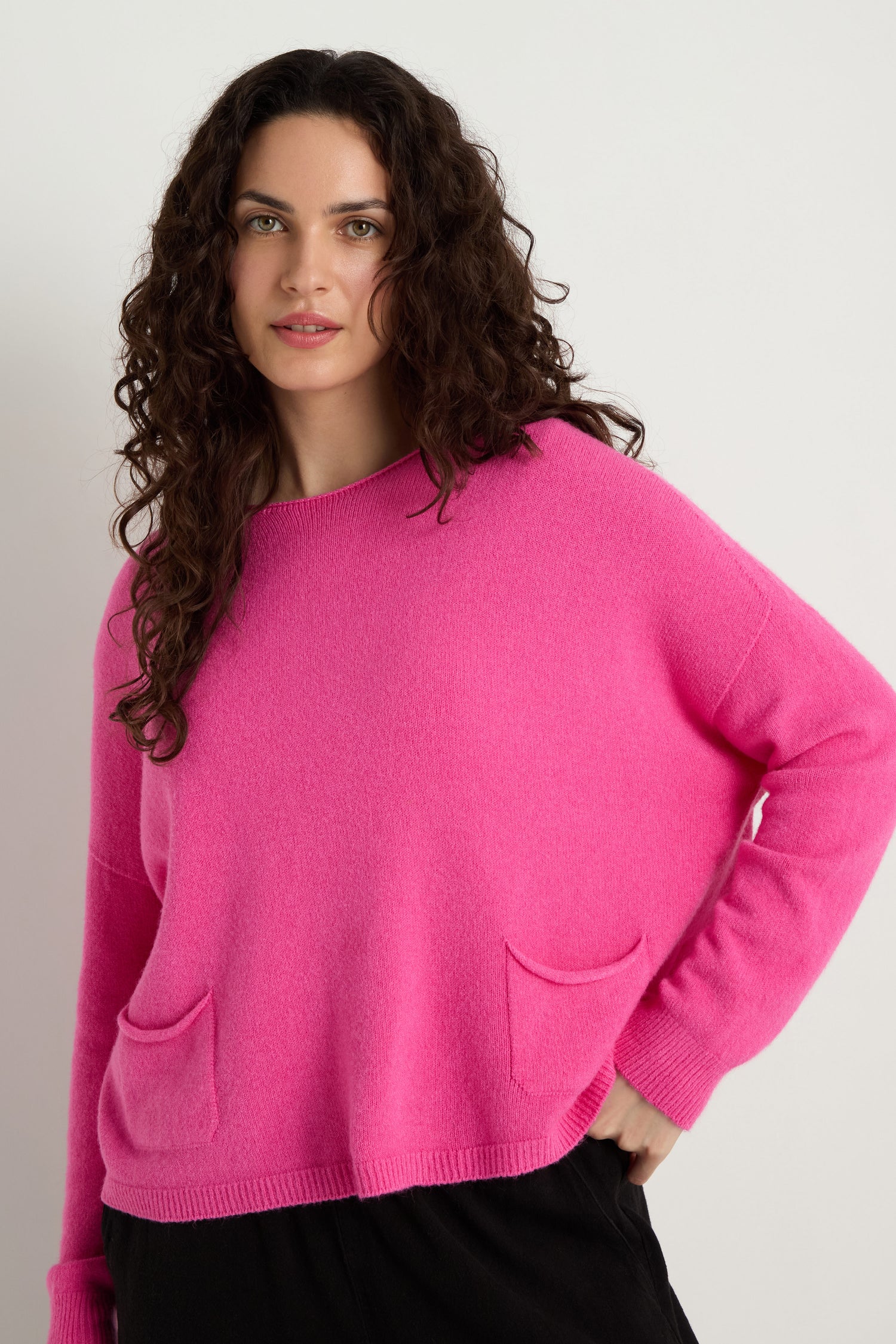 A person with long curly hair wears the Two Pocket Knit, a bright pink women's sweater with front pockets, ideal for everyday wear, standing against a plain white background.